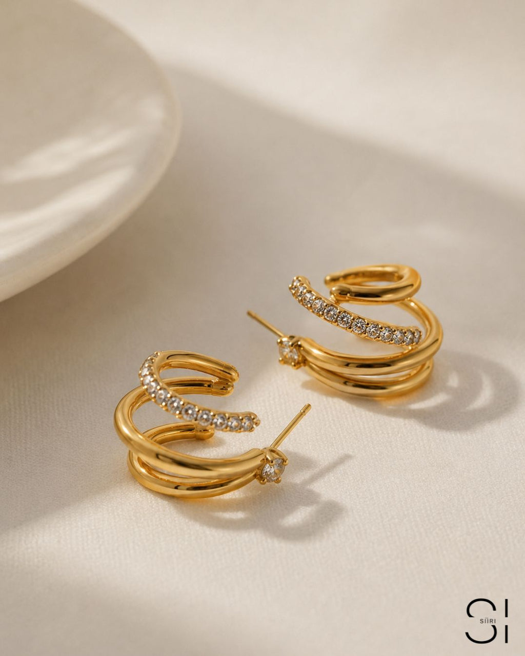 Close-up of Ciara's Diamond Overlay Studs featuring sparkling white rhinestone detailing. Gold hoop earrings with embedded stones on a beige surface