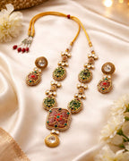 Ornate gold necklace with green and red stones on a satin surface with flowers.