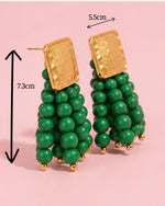 Green beaded earrings with gold accents on a pink background, showing measurements.