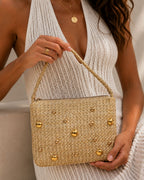 Woman holding a straw handbag with gold embellishments against a neutral background